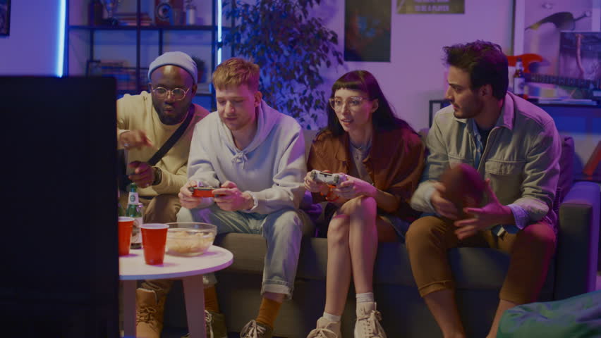 Full shot of four happy multiracial young people sitting on couch in front of TV at home in neon blue light, playing video game on console with wireless controllers, cheering and laughing Royalty-Free Stock Footage #1106583805