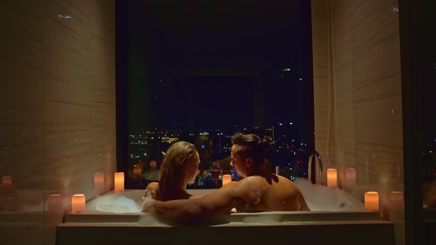 Love couple enjoys romantic honeymoon in hotel. Young married couple relax in bathtub with window view of city night. Embracing in foam, kiss hug.Honeymoon vacation romantic atmosphere in candlelight Royalty-Free Stock Footage #1106584969