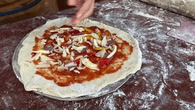 A woman wearing apron is sprinkling the dough with mozzarella cheese, sausage, peppers and vegetables toppings during a pizza cooking class session. 4k video footage