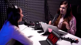Side angle view of young woman podcaster speaking to her female guest into microphone, both wearing headphones in a modern video studio 