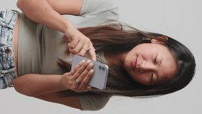 Vertical video, Happy girl in gray top scrolls on the screen of smartphone, browses social networks, receives good news, uses good mobile applications isolated on white background, studio shot