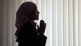 Muslim woman wearing pink hijab prays for more peaceful life in semi-darkened apartment. Religious lady asks Allah for best opportunities for family - Powered by Shutterstock - Get 15% off with code: PIKWIZARD15