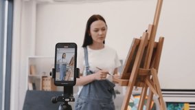 Professional female artist recording video on camera of personal smartphone during painting process for own followers. Lady in denim jumpsuit on blurred background blogging about hobbies and art.
