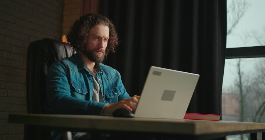Concentrated young man working on a laptop computer while sitting on a black leather chair in a stylish office. A man thinks about solving a problem and touches his faceA young freelancer works from