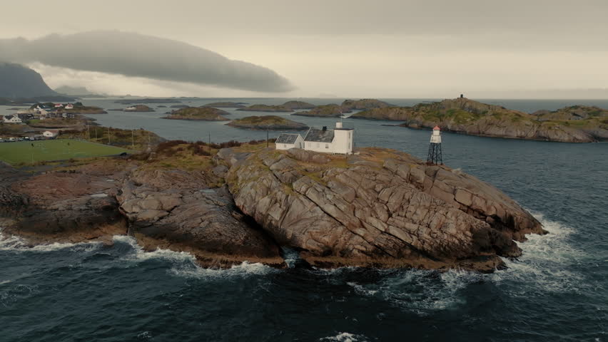 White house on the top of green rocky cliff, washed by waves in Henningsvaer commune. Aerial view.