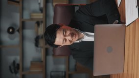 Emotional company's ceo laughing behind computer screen while resting in commodious office. Vertical video of asian executive enjoying ridiculous online clip as stress coping method during break.