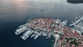 Aerial drone video flying backwards from the old Croatian town of Korcula as a ferry is leaving, revealing the sunrise and surrounding mountains and islands.