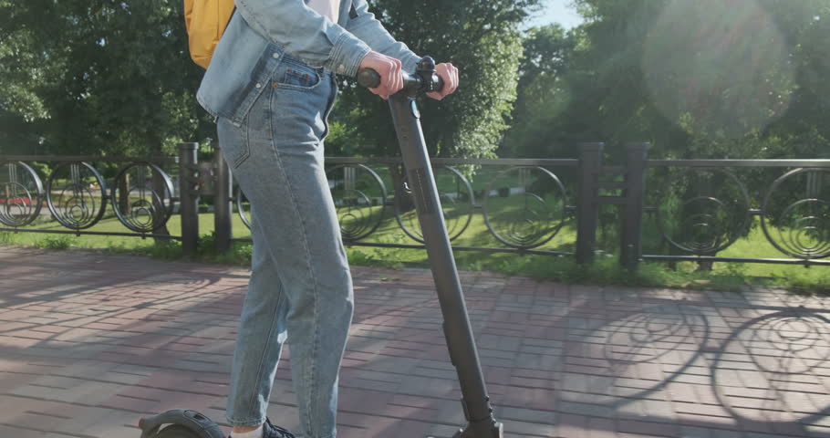Beautiful young smiling woman moves on electric scooter along city park. Camera follows girl, side view. Rough road, shaking, bumpy Royalty-Free Stock Footage #1106630497