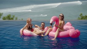 Group of friends have party swimming with pink inflatable flamingo, having fun on their holidays in pool resort, with the ocean beach on background. Young people in swimsuits laugh together - Powered by Shutterstock - Get 15% off with code: PIKWIZARD15