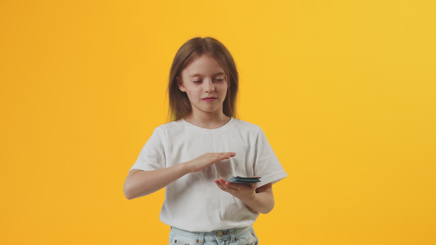 Funny Caucasian girl kid wearing white t-shirt hold fan of cash money in dollar banknotes and throws money in all directions on plain yellow background in studio