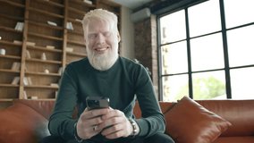 Young smiling albino man holding smartphone device texting sms message sitting at home office. Positive guy using apps playing mobile game chatting in social media surfing web on phone in modern home - Powered by Shutterstock - Get 15% off with code: PIKWIZARD15