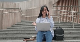 Smiling Asian young student female seats on the stairs using laptop and talking on mobile phone