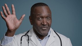 Close-up smiling african american man doctor look at camera wave hello in blue background studio happy ethnic male therapist pediatrician surgeon in white medical coat greet patient online webcam view