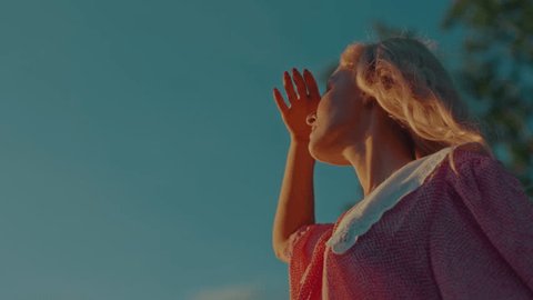 A girl in a vintage dress covers her eyes from the sun while leaning on her turquoise-colored retro car Video Stok