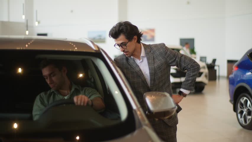Front view of stylish car dealer in business suit talking to young man client sitting in car salon. Professional salesman helping male client to choose best automobile in showroom. Royalty-Free Stock Footage #1106644593