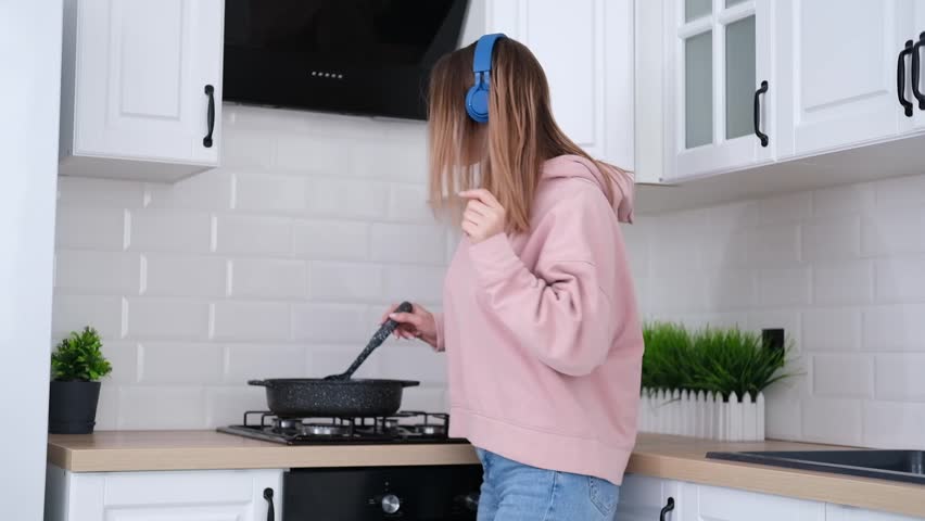 young overjoyed attractive woman wearing headphones having fun , dancing and cooking in modern light kitchen, having pleasure preparing romantic dinner alone indoors.