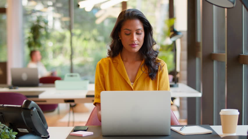 Mature businesswoman working on laptop at desk in modern office pauses to look out of the window and smile - shot in slow motion