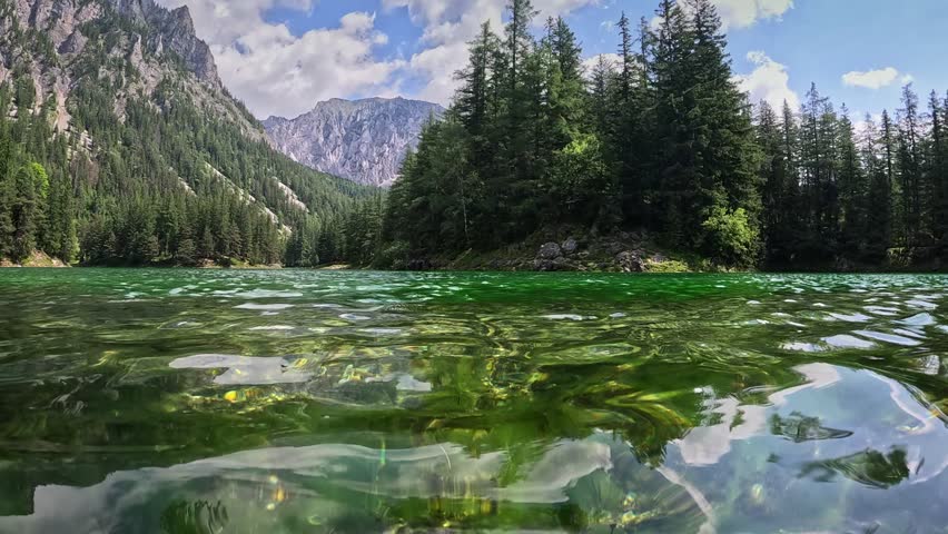 Half underwater shot of mountain lake with clear water in the Alps of Austria. Camera dive into the lake, rocky mountains and forest in the background. Alpine nature, Gruener See