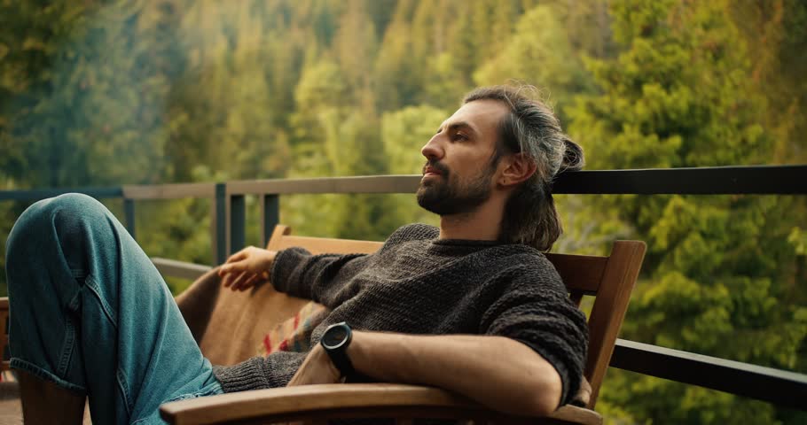 A young brunette man is resting, sitting on a sofa against the backdrop of mountains and coniferous forest