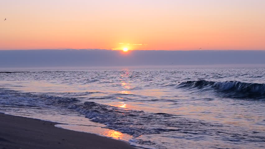 Beautiful sunrise on sea coast seaside. Sun rising from behind clouds in sky above horizon line on blue sea with sea waves rolling on sandy beach. Beautiful seascape sea landscape. Natural background