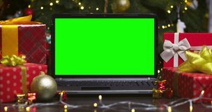 Laptop with green screen surrounded by gifts on background of Christmas tree