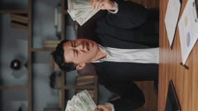 Vertical video of fortunate businessman playing with dollar billes before throwing them in air of office room. Enthusiastic asian entrepreneur dancing in rain out of banknotes during working day.