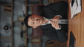 Portrait of friendly asian businessman waving hand with greeting while posing at camera in workplace. Vertical video of positive employee inviting for online webinar on marketing strategies in office.