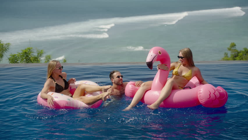 Diverse group of young friends having fun in infinity resort pool with pink inflatable flamingo. Cool party with water splashing and swimming, chilling with the ocean beach view | Shutterstock HD Video #1106674557