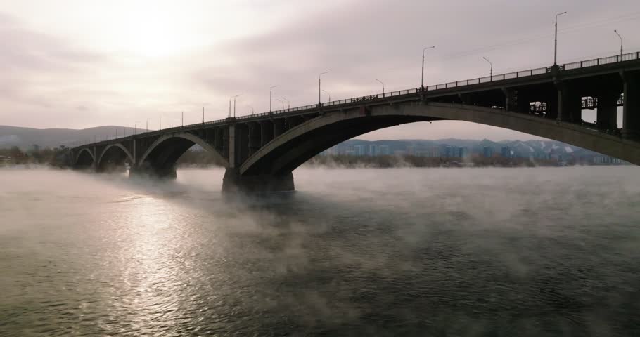 POV aerial view from a copter, flying under the municipal bridge of Krasnoyarsk, over the Yenisei River, fog is rising over the water, winter time Royalty-Free Stock Footage #1106687411