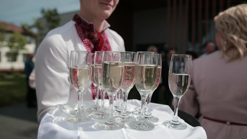 Waitress is holding tray with champagne glasses in her hand. The waiter serves people at the restaurant. Alcohol drinks in wine glasses. Night party, holidays event outdoor. People drinking.