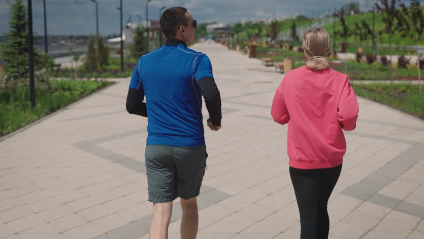 Sports couple on a daily run in the city park. Bright summer day and landscaped park area. Convenient urban environment for sports and recreation. Royalty-Free Stock Footage #1106691435