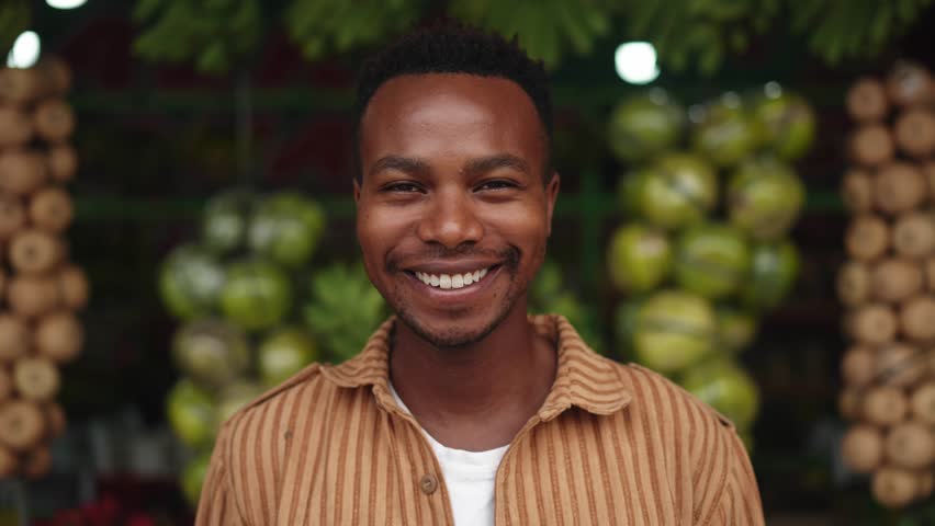 Portrait of Young Black Man Looking at Camera on Fruit Market Background. One Smiling Face and Beautiful Eyes of Happy Male Person Outdoors. Optimism of African American Boy Standing on City Street 4k