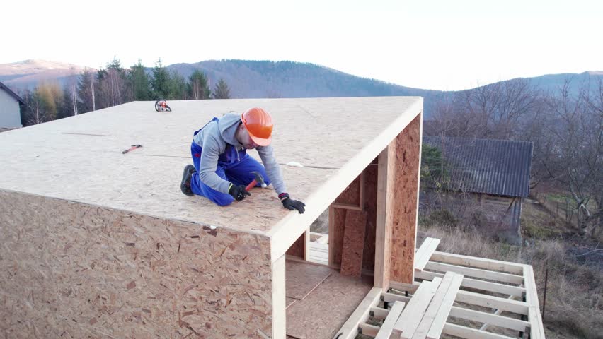 Carpenter hammering nail into OSB panel on the roof top of future cottage. Man worker building wooden frame house. Carpentry and construction concept.