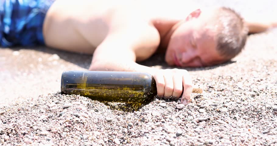 Drunk young man with bottle of wine in his hand lying on seashore 4k movie