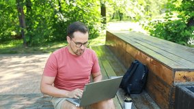 A man is working on a laptop, typing text on the keyboard, sitting on a bench outdoors in the park on a summer day. Remote work or study