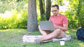 A man works remotely at a laptop while sitting on a plaid on the grass in a park on a summer day. Remote work or online training