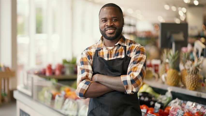 Portrait of a Black-skinned man in a plaid shirt and black apron folded his arms over his chest and looks at the camera in a supermarket Royalty-Free Stock Footage #1106713113
