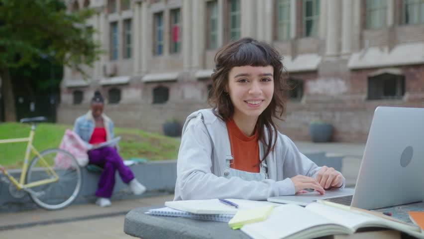 Young pretty college girl sitting with laptop and workbooks outdoors on campus and happily smiling at camera. Medium shot, video portrait
