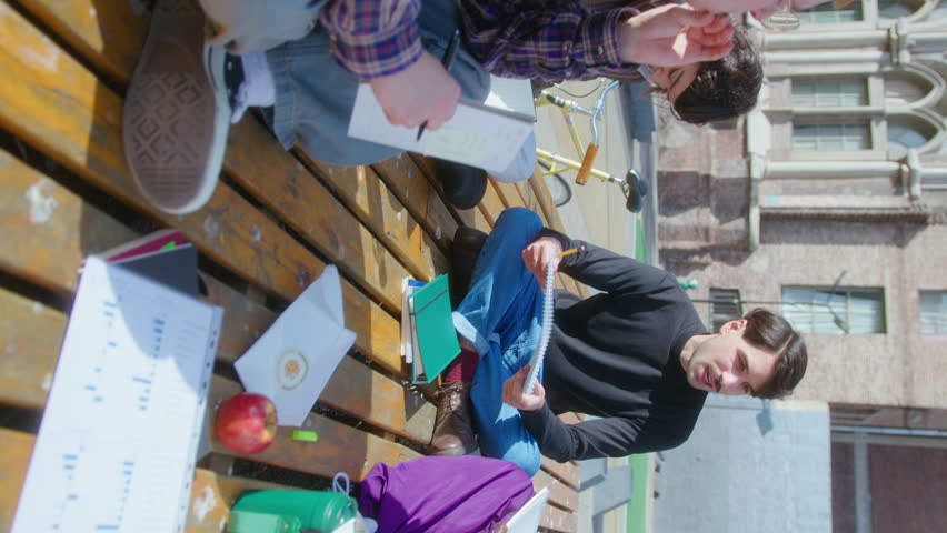 Young multi-ethnic group mates making notes in workbooks, discussing college project, sitting on a bench with snacks and drinks outdoors on campus. Vertical video Royalty-Free Stock Footage #1106713803