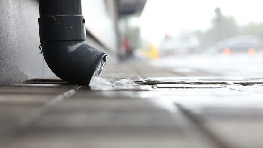 Rainwater flows from a drainpipe onto the sidewalk, people walk under an umbrella in the blurred background. Rainy weather. Royalty-Free Stock Footage #1106720479