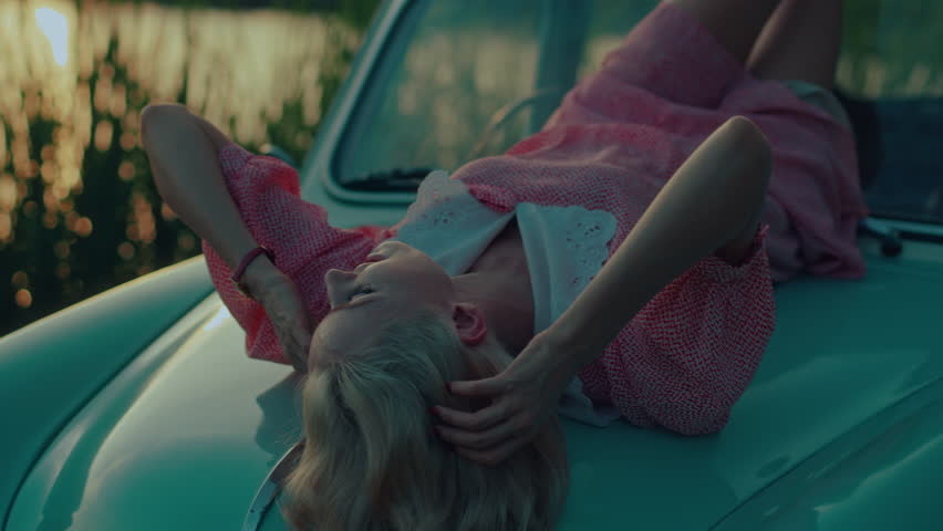 A blonde in a pink vintage dress lies on the hood of a turquoise-colored retro car