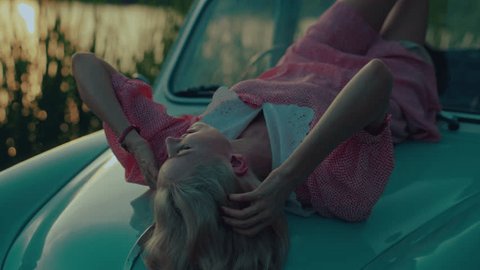 A blonde in a pink vintage dress lies on the hood of a turquoise-colored retro car Stockvideo