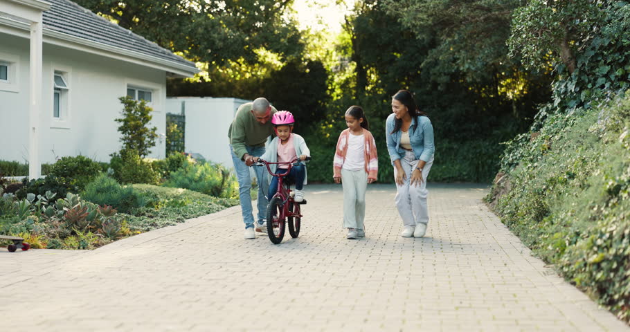Family, parents and girl with learning, bicycle and sister with support, teaching and coaching for skill in driveway. Father, mother and daughter kids with bike, helping and applause in home backyard