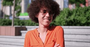Happy smiling young African American woman or student with curly hair posing standing outdoor on sunny city street looking at camera
