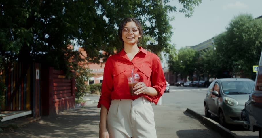 A young woman in casual clothes smiles, and looks at the camera drunk lemonade from a disposable cup with a straw standing on a city street