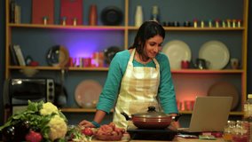 An Indian woman joyfully cooks in the kitchen, chatting on a laptop, showing happiness.
