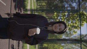 Attractive smiling girl in a brown sweatshirt listens to music on headphones and drinks takeaway coffee from a paper cup on the sports ground. A girl on the sports ground drinks takeaway coffee.