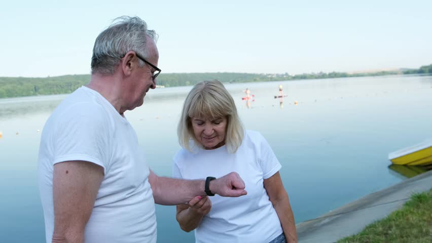 Seniors talking by the lake using a smart watch. Sports training after retirement