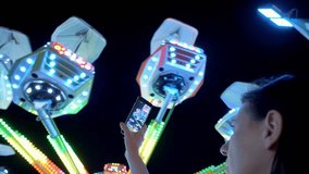 Woman hands are holding smartphone and taking photo or shooting video. Carousel with lights. Incredible colorful flashing light of vintage carousel.Popular Chair Swing Rides in Amusement Park