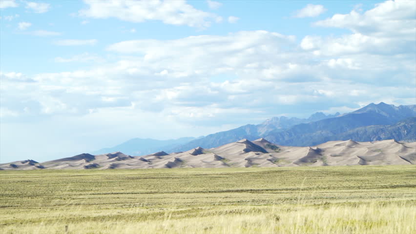 Stunning late summer early fall view of The Great Sand Dunes National Park Colorado Rockies mountain sandy 14er peaks crisp golden yellow tall grass wind clouds blue sky mid day cinematic car motion